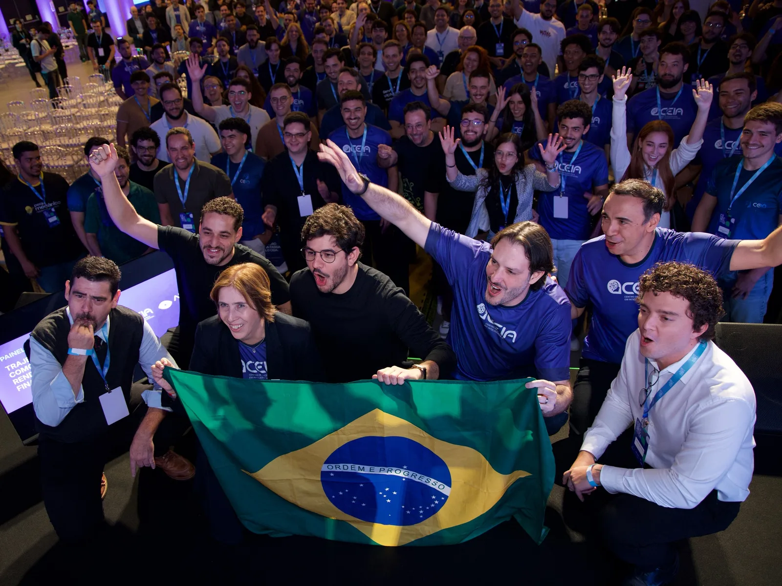 Pesquisadores do CEIA erguem a bandeira do Brasil diante de um auditório lotado.
